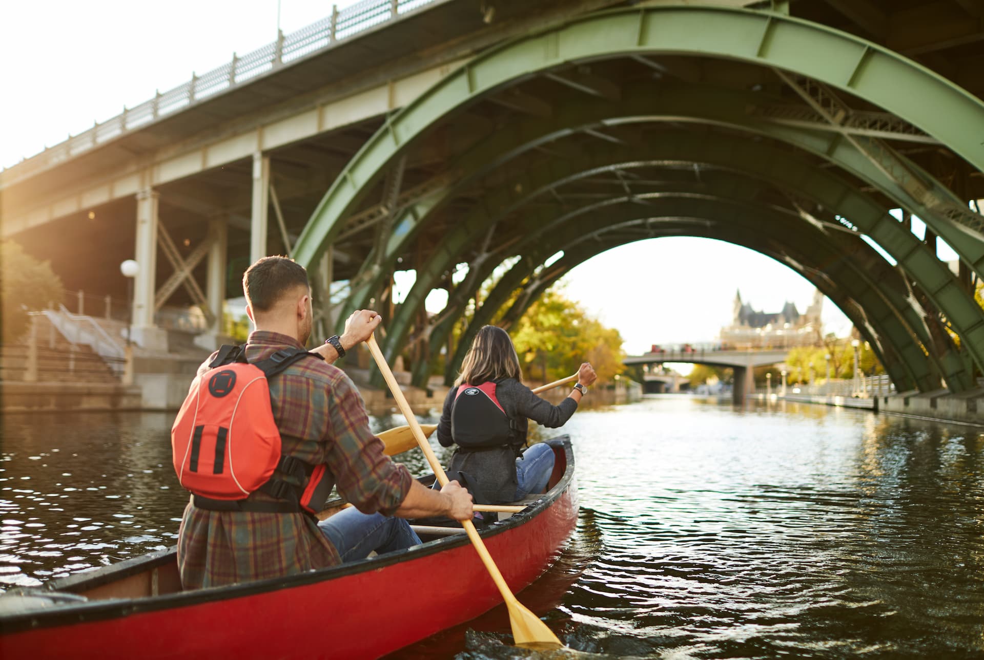 Two people paddle a red canoe under a green iron bridge on a calm urban waterway at sunset.