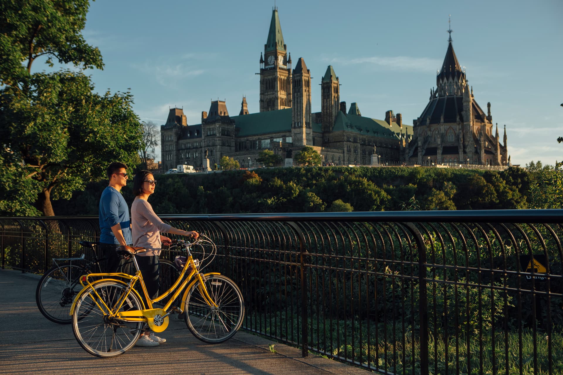 Two people with bicycles stand by a railing, overlooking Ottawa’s Parliament Buildings on a clear day.