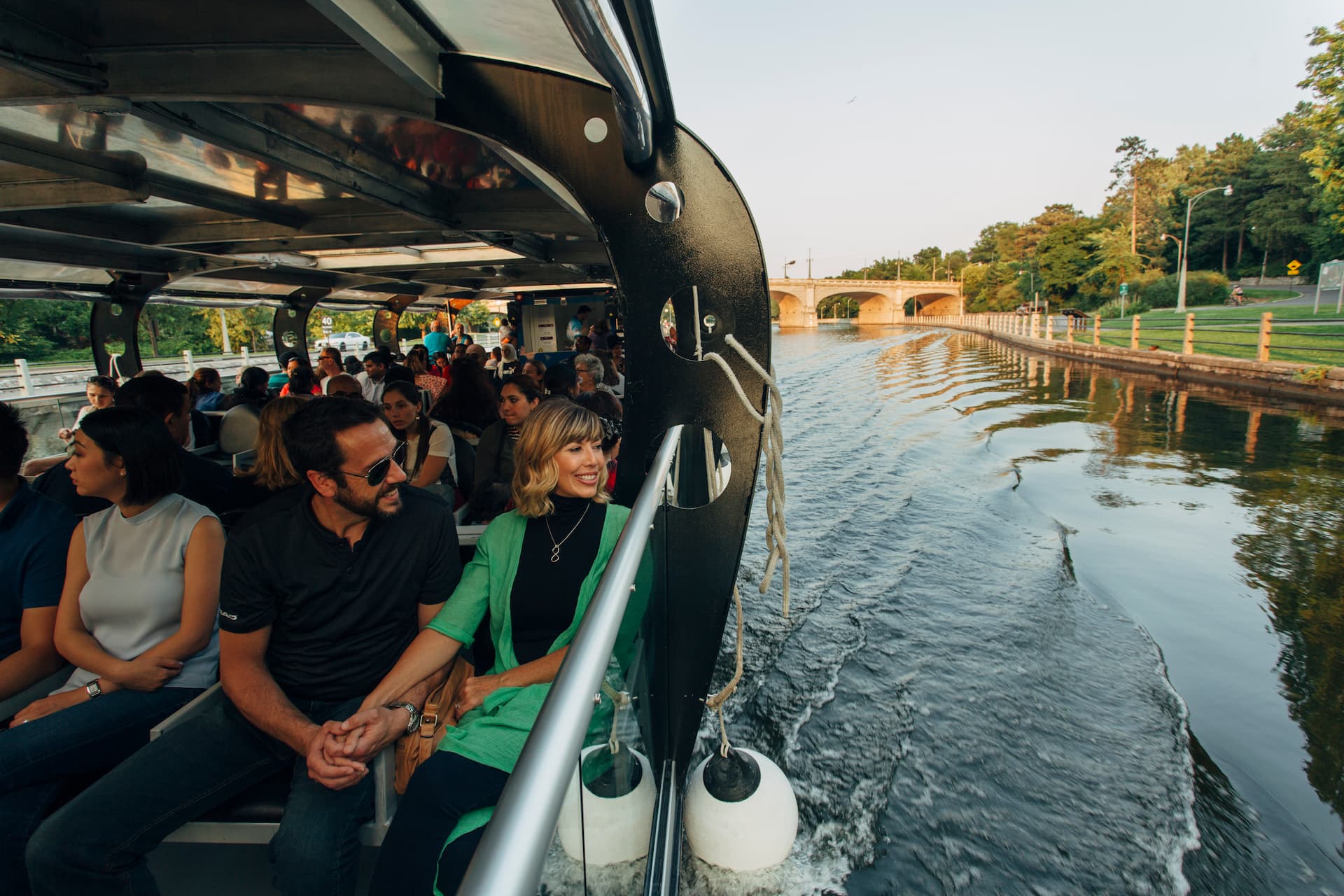 A group of people ride on a riverboat, passing under a bridge with trees and a walkway along the water.