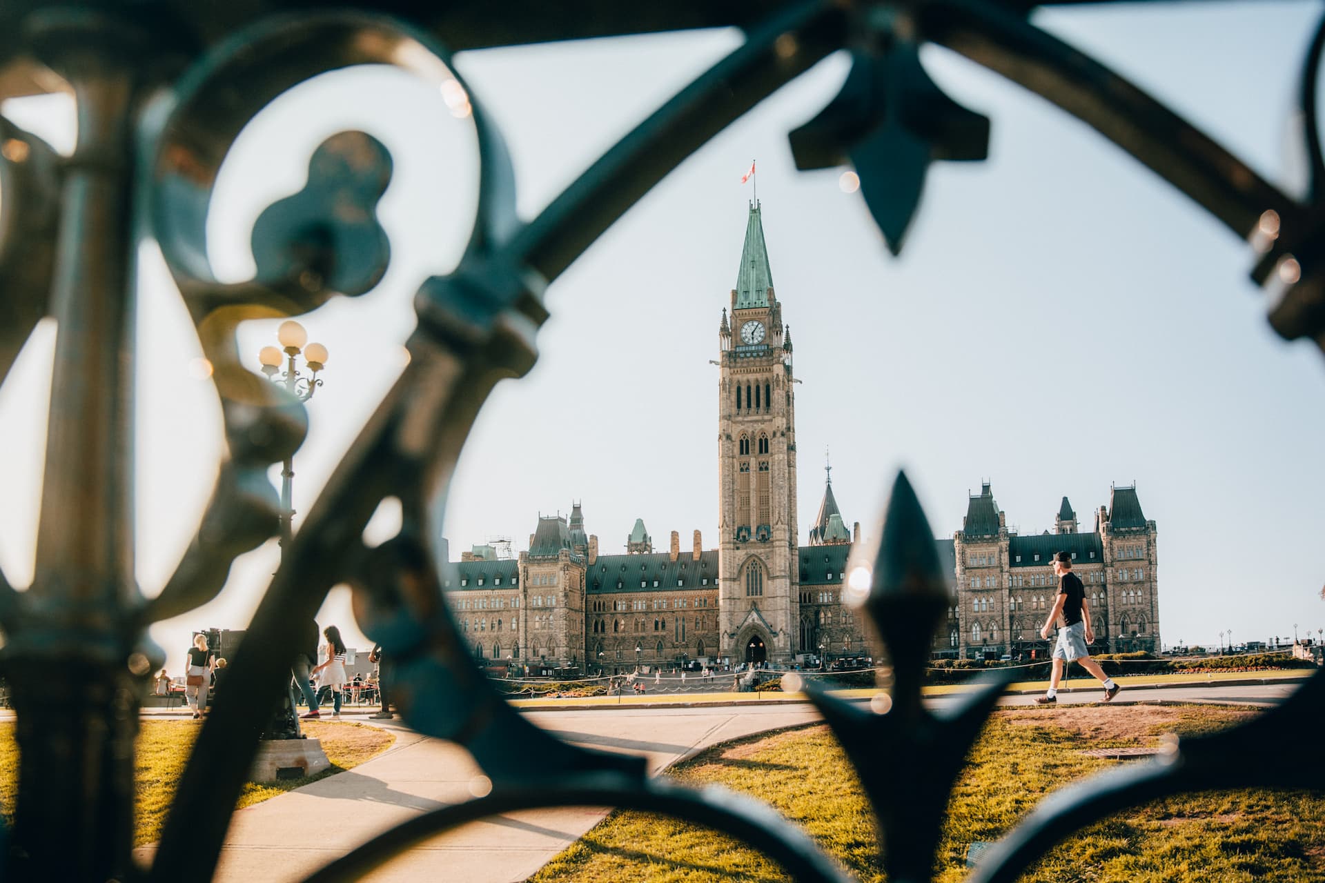 View of a large historic building with a central clock tower, seen through ornate metalwork, with people walking outside.