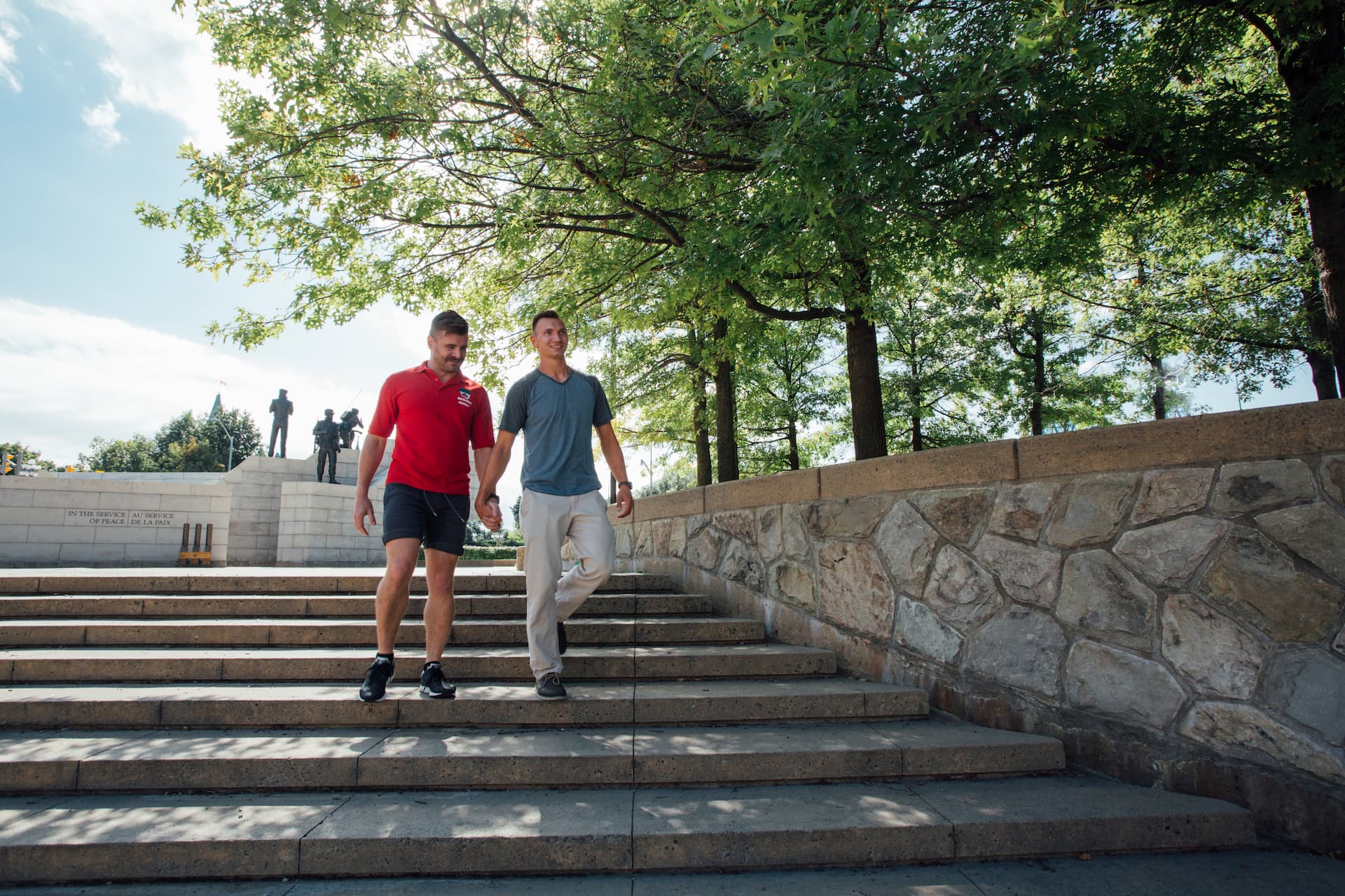 Two men walk down outdoor steps near a stone wall, with trees and a statue visible in the background.