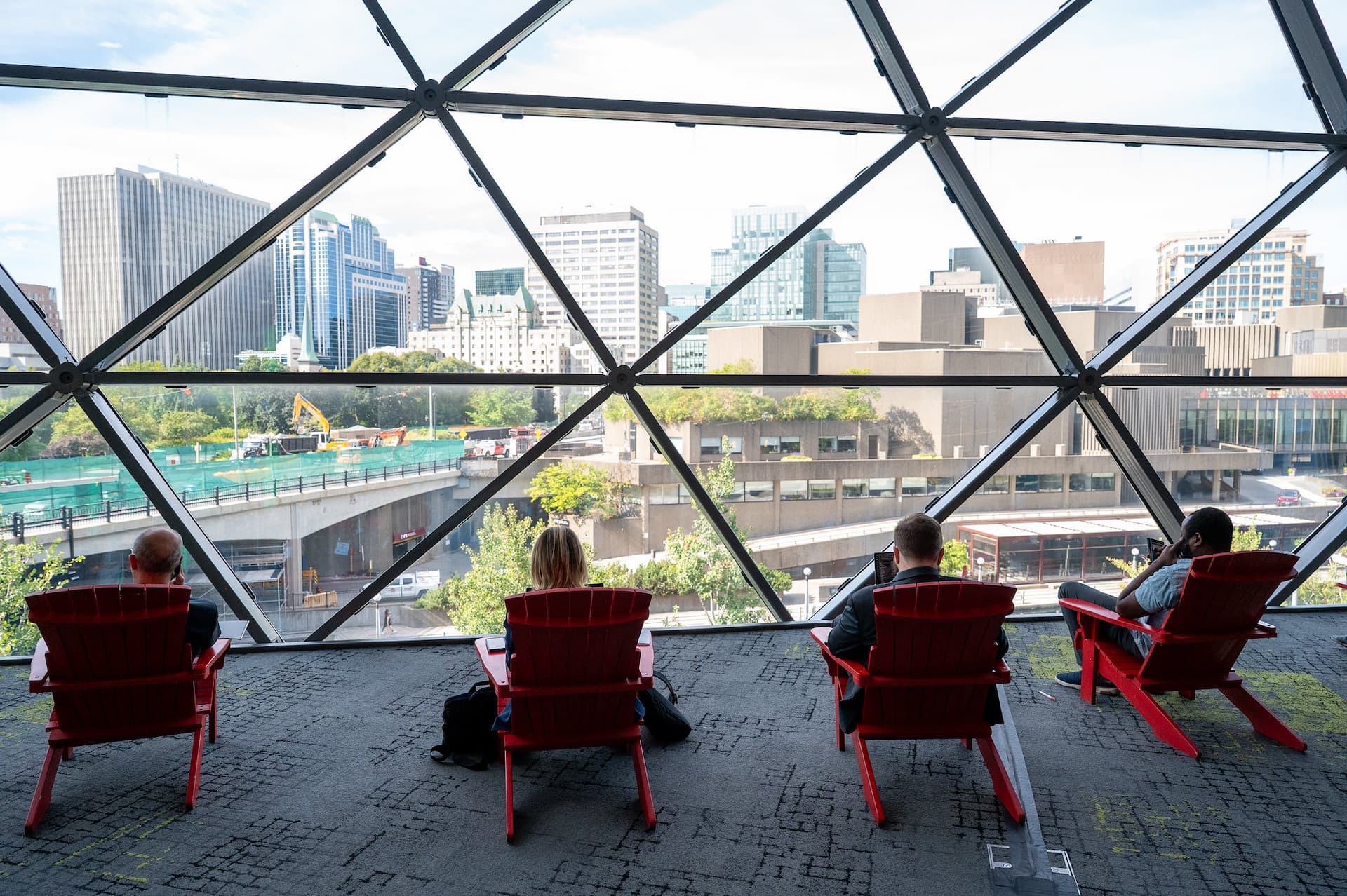 Four people sit in red chairs facing large windows with a cityscape view of high-rise buildings and a construction site.