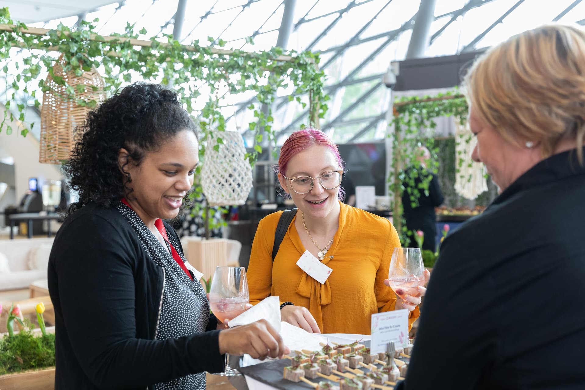 Three women stand at a table with drinks, smiling and selecting appetizers at an indoor event.